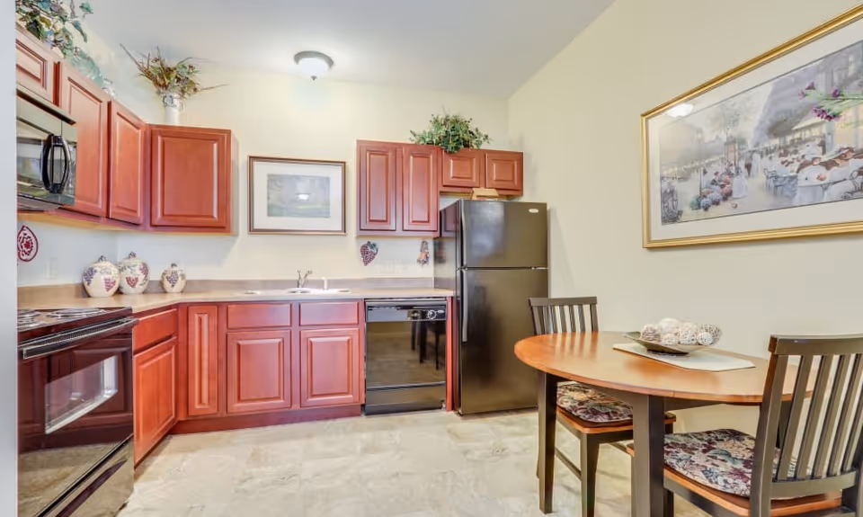 A kitchen area with wooden cabinets, a black refrigerator, a black dishwasher, a black stove, and a microwave. There is a round wooden dining table with two chairs that have floral cushions. The walls are light-colored and decorated with framed artwork and greenery on top of the cabinets.