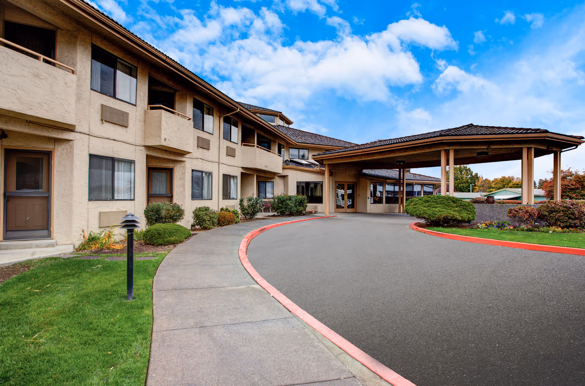 Exterior view of Solista Walla Walla facility showing a two-story building with beige stucco walls, multiple windows, and small balconies. There is a covered entrance with a driveway and landscaped bushes and grass under a partly cloudy blue sky.
