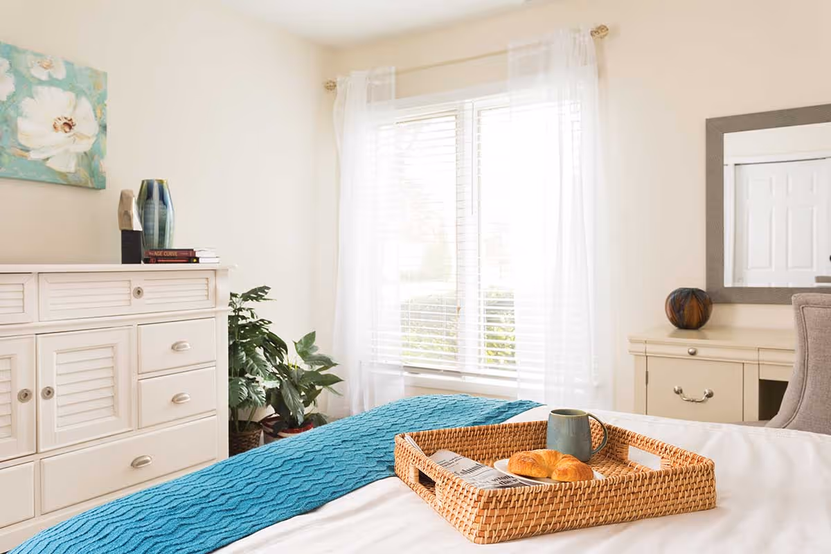Bright bedroom with a bed featuring a wicker tray holding a mug and pastry, white dressers, a window with sheer curtains, and a potted plant.