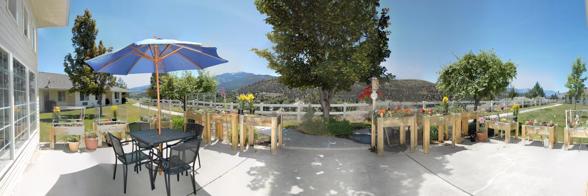 Outdoor patio area with a black metal table and four chairs under a blue umbrella. Several wooden planter boxes with flowers and plants are arranged along the concrete patio. Trees and a white fence surround the area, with hills and a clear blue sky in the background.