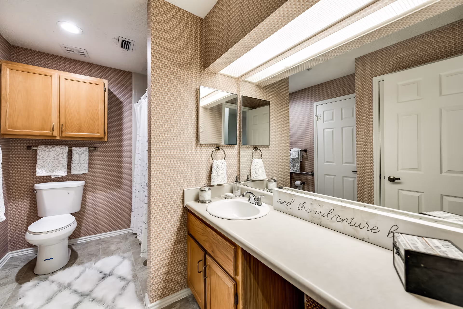 A bathroom with a white toilet, wooden cabinets above the toilet, and a countertop with a sink. There are two mirrors above the sink, a towel ring with a white towel, and a decorative sign on the countertop. The walls have a patterned wallpaper, and there is a white door and a shower curtain visible in the background. A fluffy white rug is on the tiled floor.