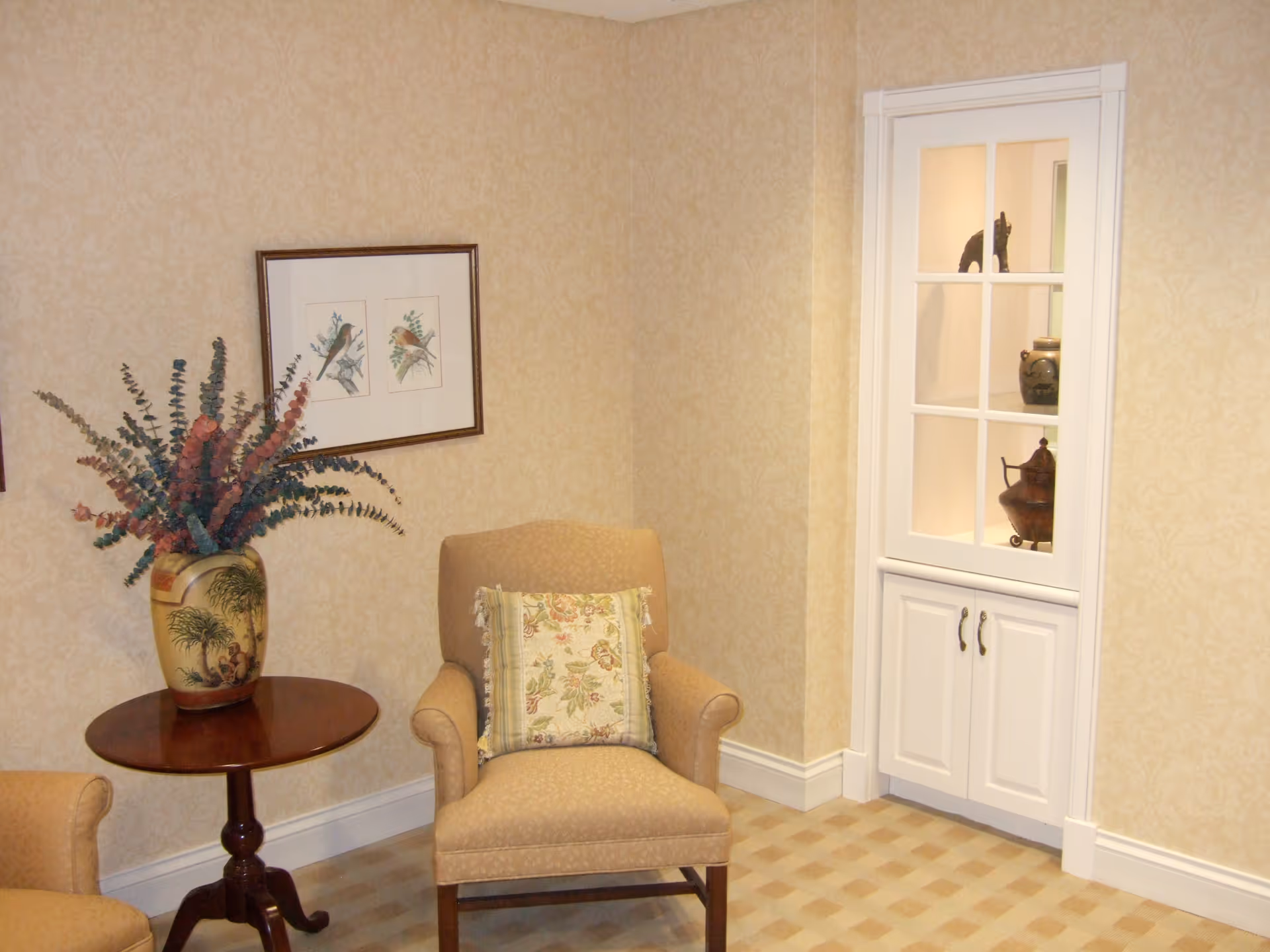 A cozy corner of a room with beige patterned wallpaper and carpet. There is a cushioned armchair with a floral pillow, a round wooden side table holding a decorative vase with dried flowers, and a built-in white cabinet with glass doors displaying pottery and a small sculpture.