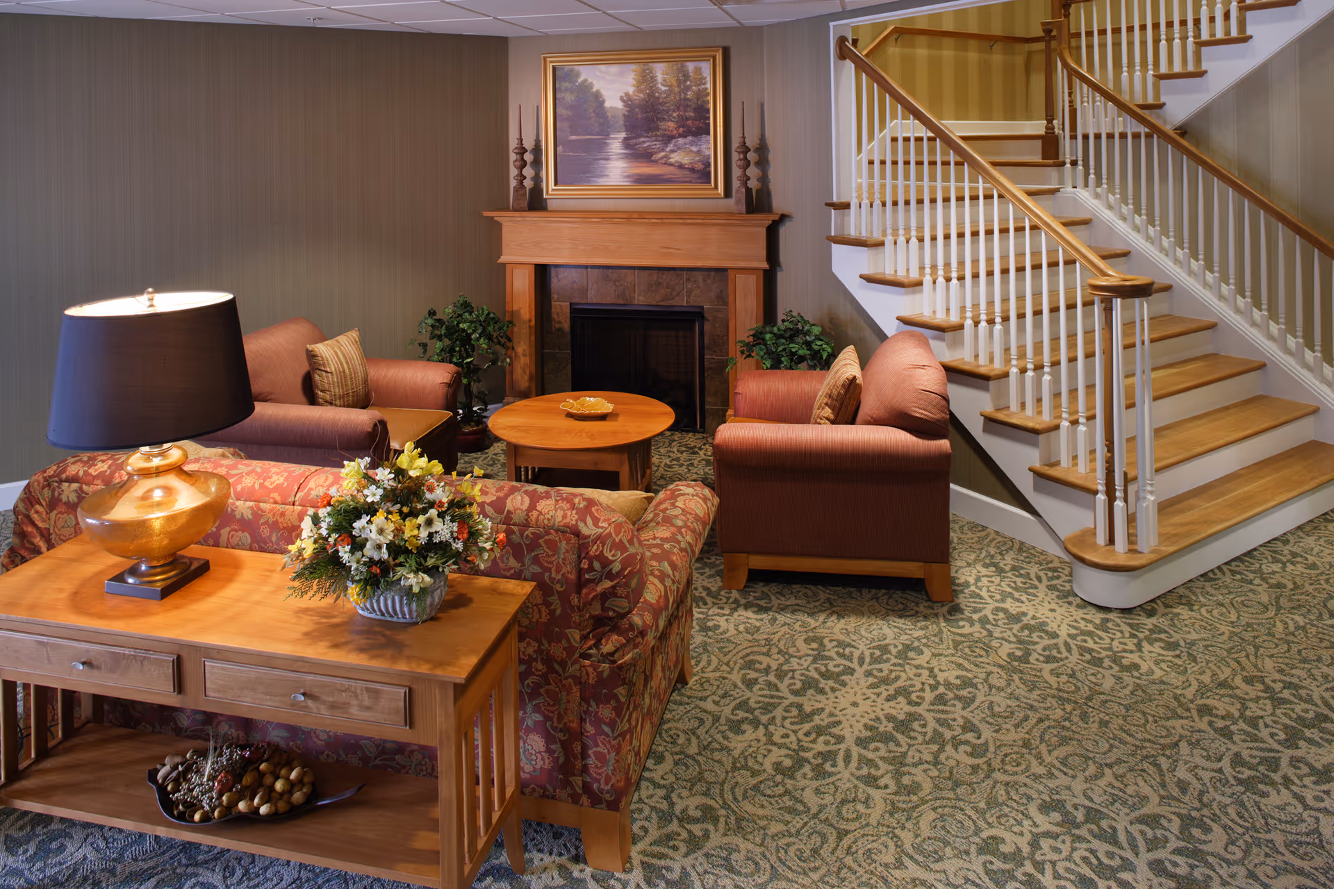 A cozy living room area with patterned carpet, a floral sofa, two armchairs, a wooden coffee table, and a wooden side table with a lamp and a flower arrangement. There is a fireplace with a framed landscape painting above it, flanked by two candlesticks. A staircase with wooden steps and white railings is visible on the right side.