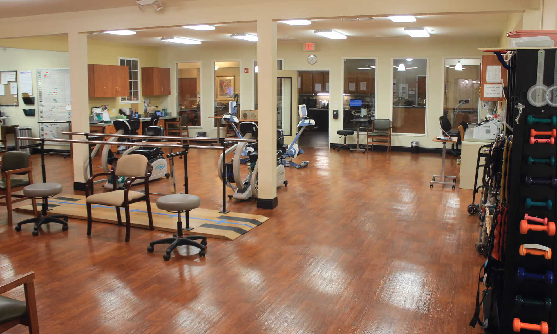 A spacious indoor rehabilitation/exercise room with parallel bars, chairs, exercise machines, and a rack of colorful dumbbells.