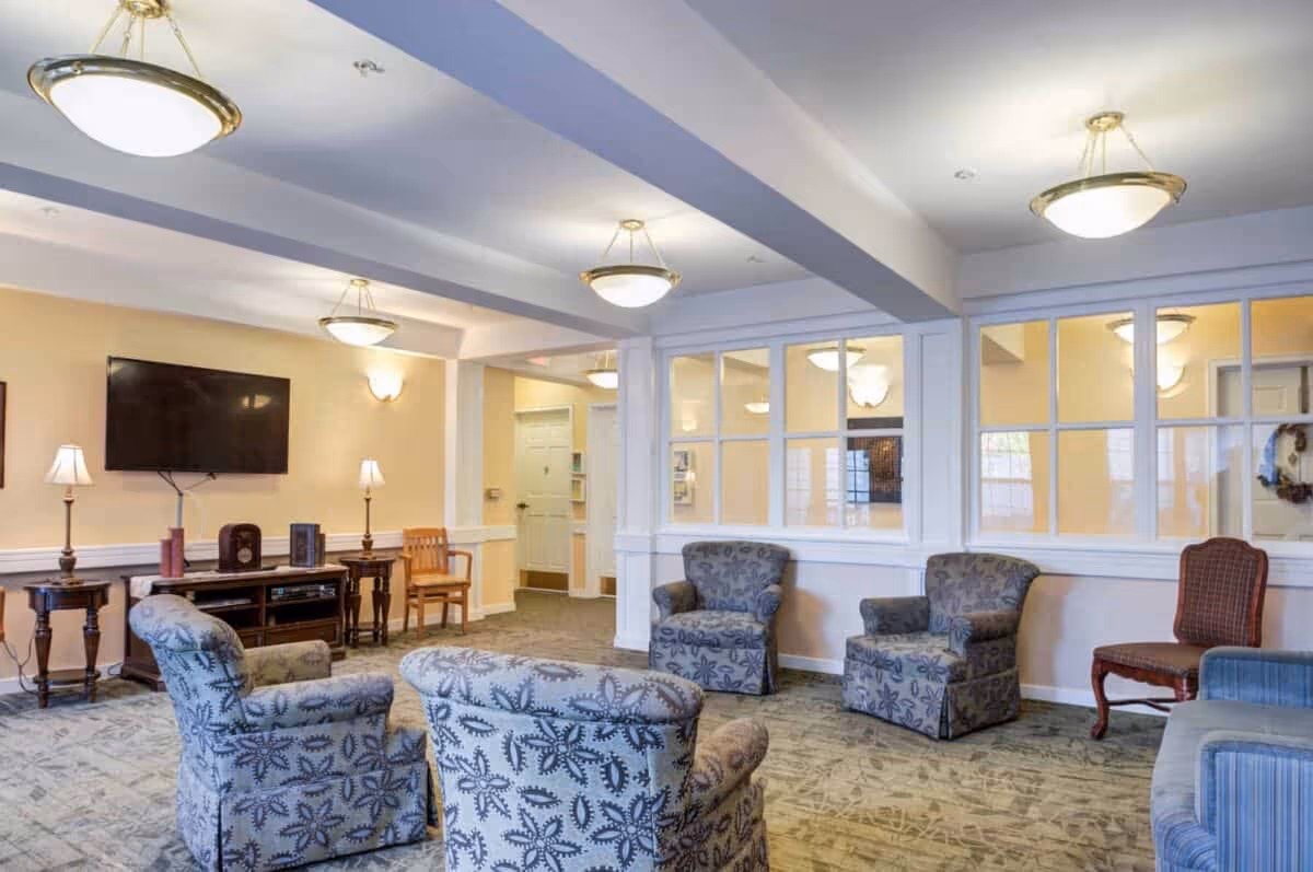 A cozy living room area in a senior living facility with patterned armchairs arranged around the room, a flat-screen TV mounted on the wall, wooden side tables with lamps, and a carpeted floor. The walls are painted a soft beige, and there are large windows with white frames separating this room from an adjacent space.
