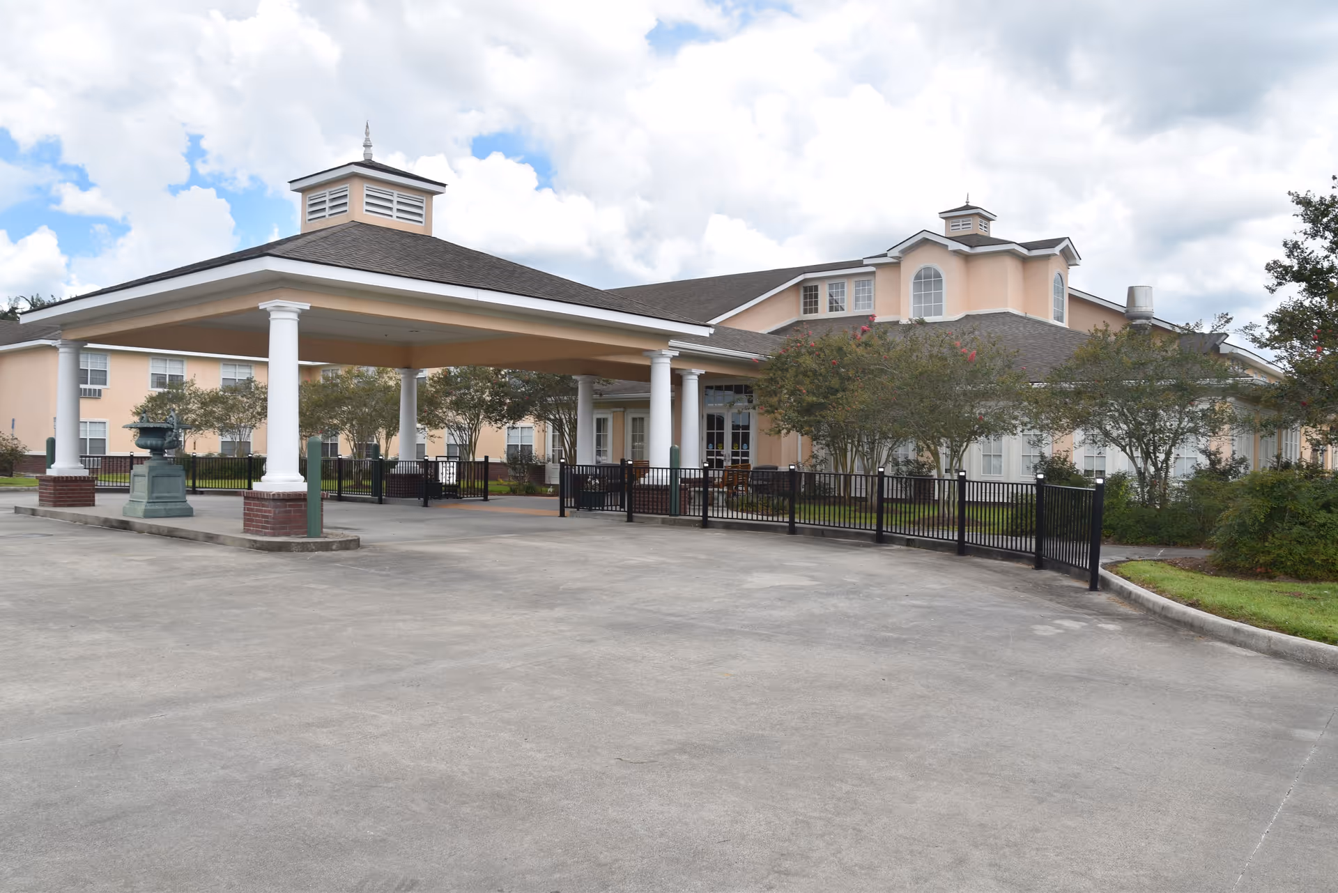 Front exterior of a senior living building with a covered porte-cochere supported by white columns, a paved driveway, landscaping, and a cloudy sky.
