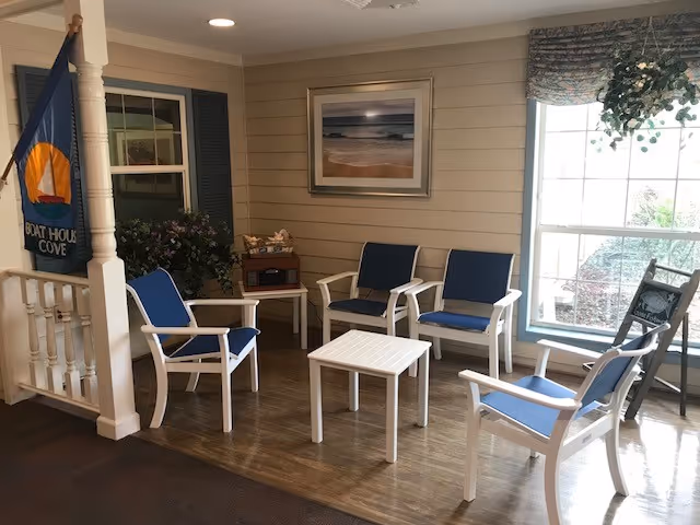 A cozy seating area with five white chairs featuring blue cushions arranged around a small white table. The room has beige shiplap walls, a large window with floral valance, a framed beach photograph on the wall, a flag with a boat house logo, and some potted plants. The floor is a combination of wood and carpet.