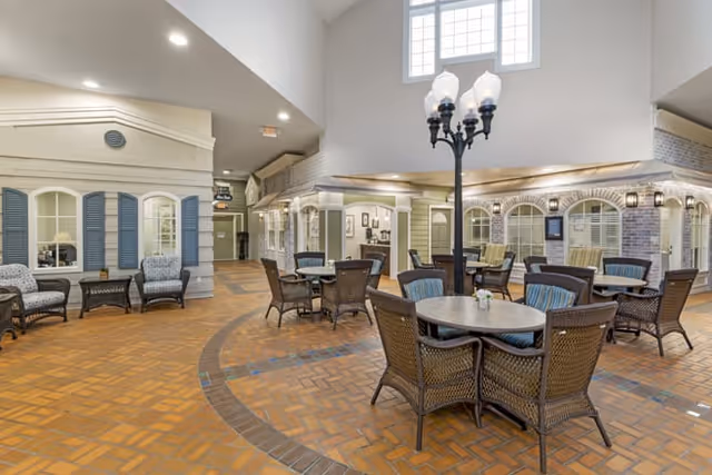 Indoor common area with brick flooring and multiple round tables surrounded by wicker chairs. The space features a high ceiling with large windows, decorative streetlamp-style lighting, and walls designed to resemble the exterior of houses with windows and shutters. Comfortable armchairs are placed along the walls.