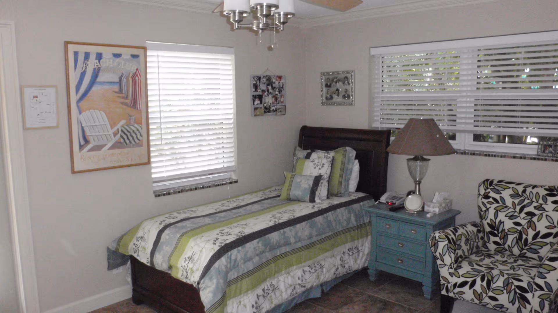 Single-occupancy bedroom with a twin bed, blue nightstand and lamp, patterned armchair, wall art, and windows with blinds.