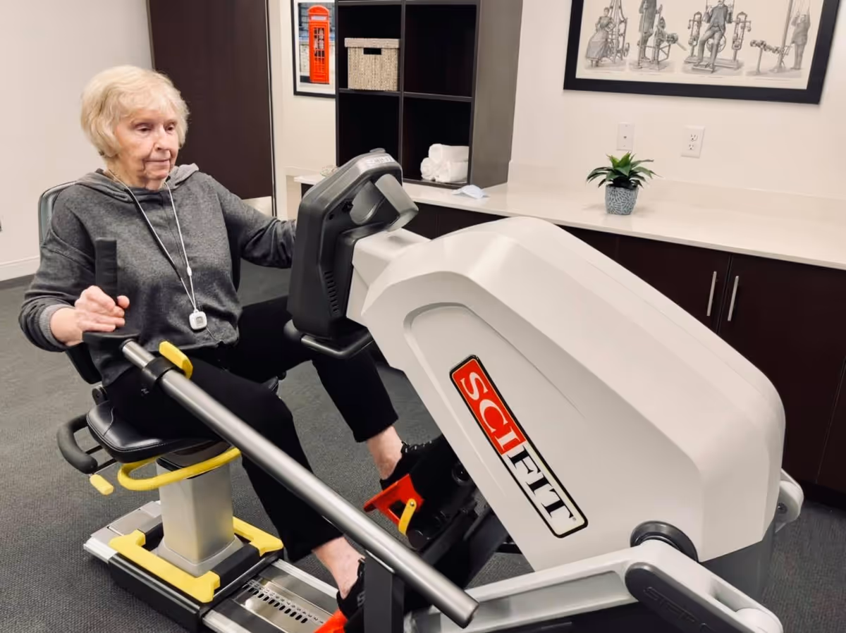 An elderly woman using a SciFit exercise machine in a fitness room with cabinets, a countertop, a small potted plant, and framed artwork on the wall.