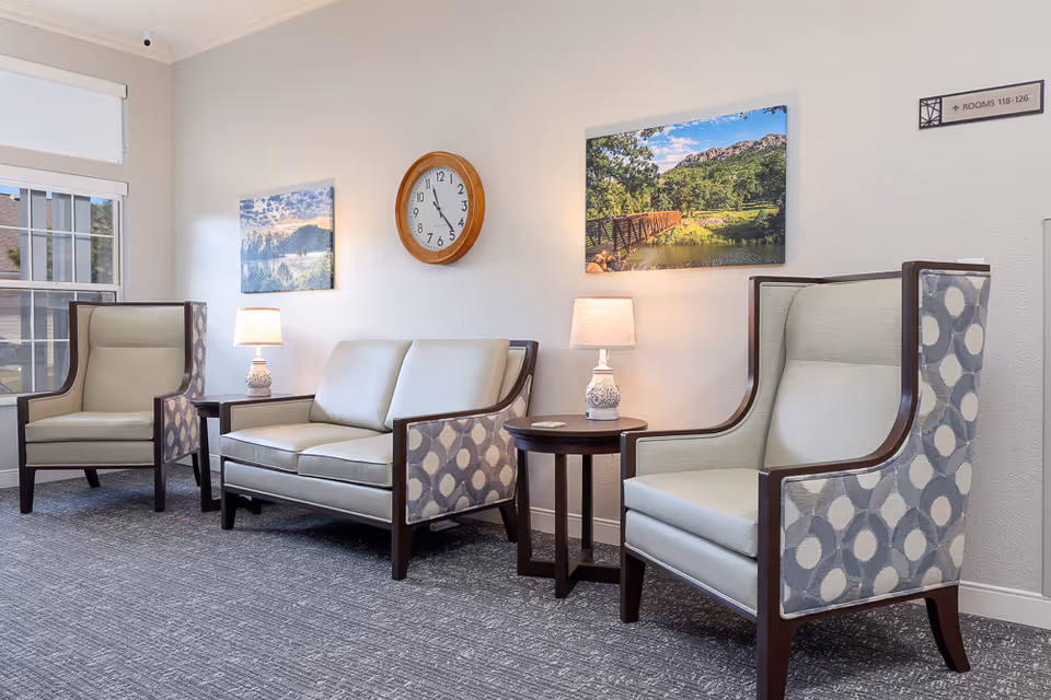 A bright and clean sitting area with two patterned armchairs and a matching loveseat arranged around two small round wooden tables with lamps. The walls are white with two landscape paintings and a round wooden clock showing the time as 11:55. A window with a white blind lets in natural light, and a sign on the wall indicates rooms 118-126.