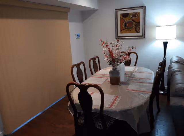 Dining room with a rectangular table covered with a tablecloth and six wooden chairs around it. A vase with flowers is placed in the center of the table along with a paper towel holder. There is a floor lamp turned on near a brown leather sofa, and a framed abstract painting hangs on the wall. A large window or sliding door with closed beige blinds is visible on the left side.