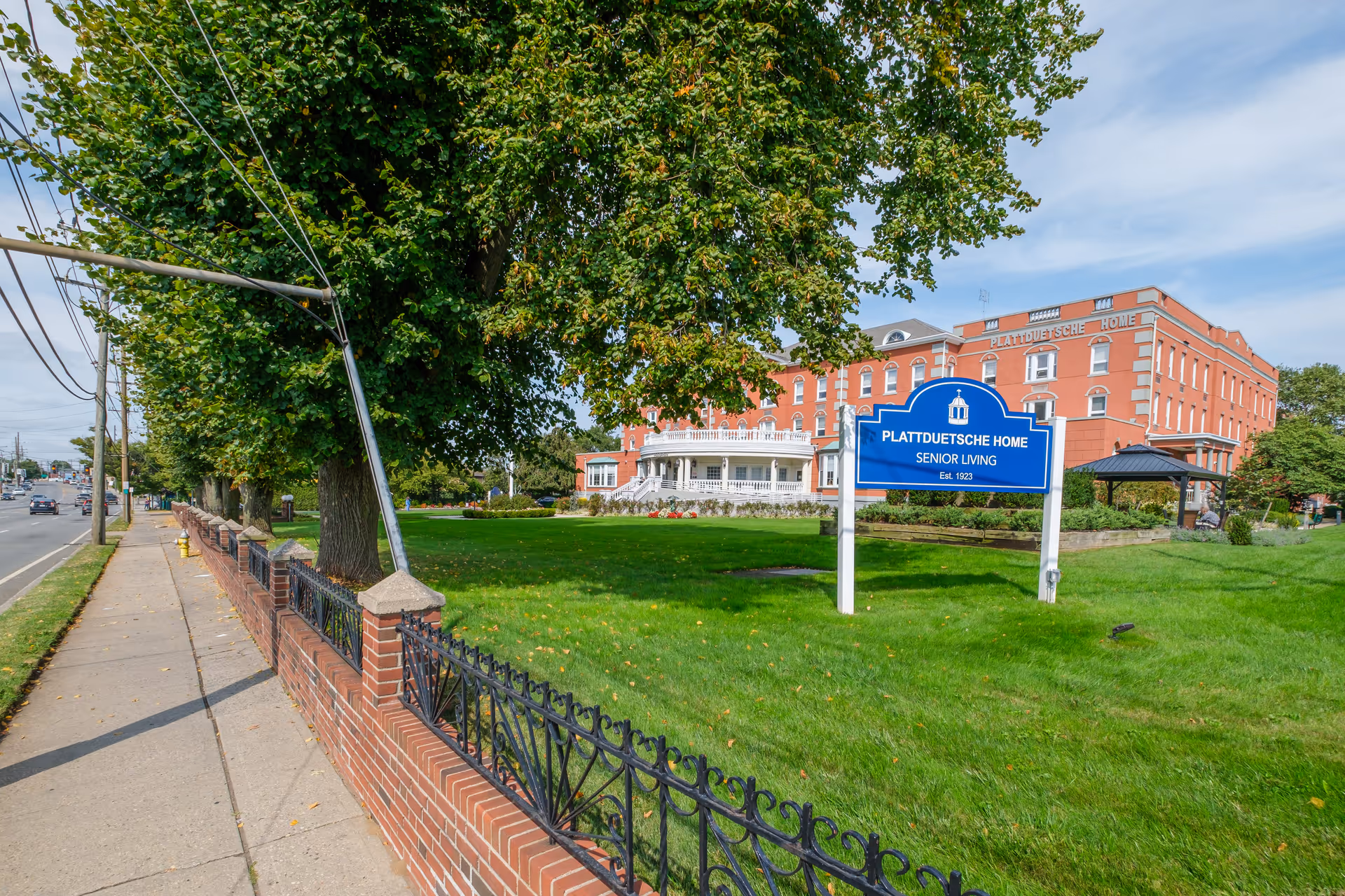 Exterior view of Plattduetsche Home, a senior living facility, showing a large red brick building with white trim. In front of the building is a well-maintained green lawn with a blue sign that reads 'Plattduetsche Home Senior Living Est. 1923'. A sidewalk and a brick and wrought iron fence run along the left side of the image, with trees lining the property. The sky is partly cloudy.