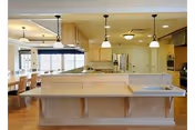 A spacious, modern kitchen with light wood cabinetry and a large island countertop. The kitchen features multiple pendant lights hanging from the ceiling, a built-in oven, a refrigerator, and a sink on the island. In the background, there is a dining area with a table and chairs near large windows allowing natural light to fill the space.