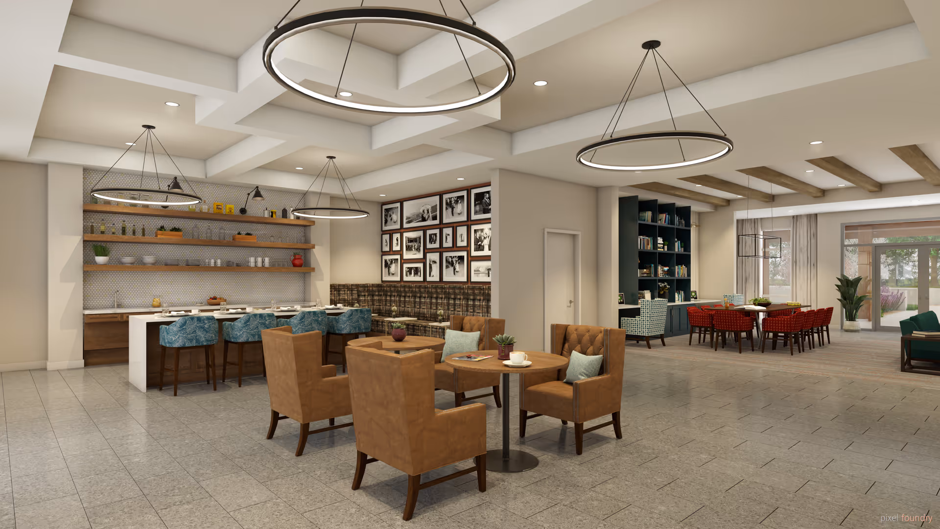 A spacious and modern common area in a senior living facility featuring a seating area with four brown armchairs around a small round table, a kitchen island with blue bar stools, and a dining area with red chairs. The room has large circular pendant lights, a bookshelf, framed black and white photos on the wall, and large windows letting in natural light.