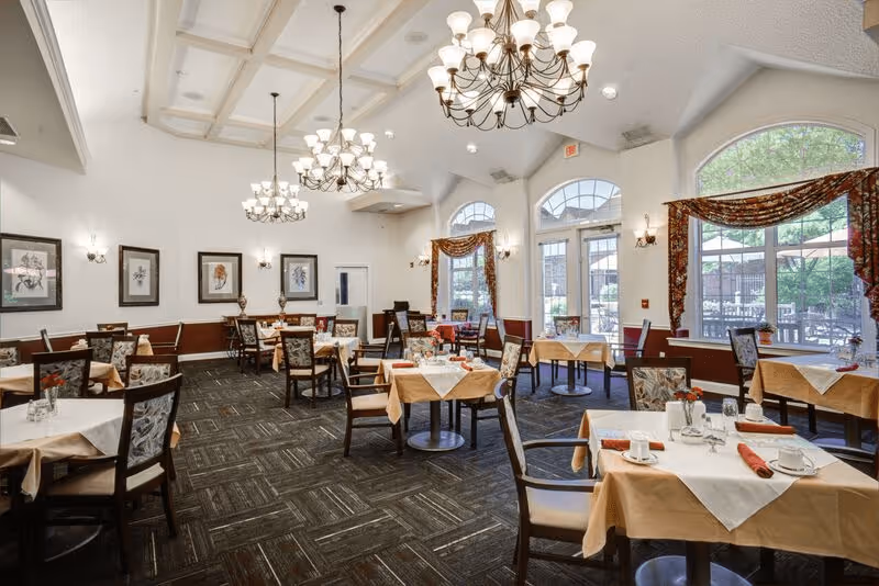 A bright and spacious dining room with multiple tables covered in beige tablecloths and white overlays, each set with cups, glasses, and red napkins. The room features large windows with patterned curtains, elegant chandeliers hanging from a coffered ceiling, framed artwork on the walls, and a carpeted floor.