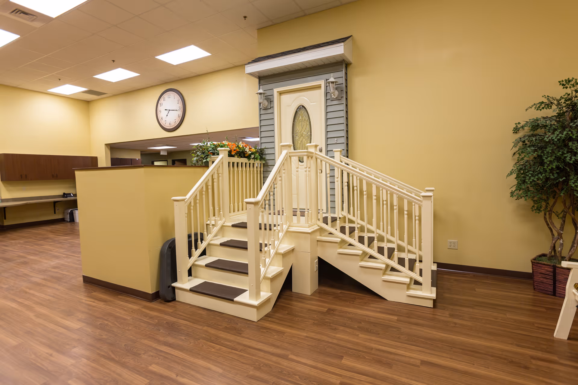 An indoor room with wooden flooring and yellow walls featuring a small staircase with handrails leading up to a white door with decorative glass. A clock is mounted on the wall above a half partition, and there are plants and cabinets in the background.