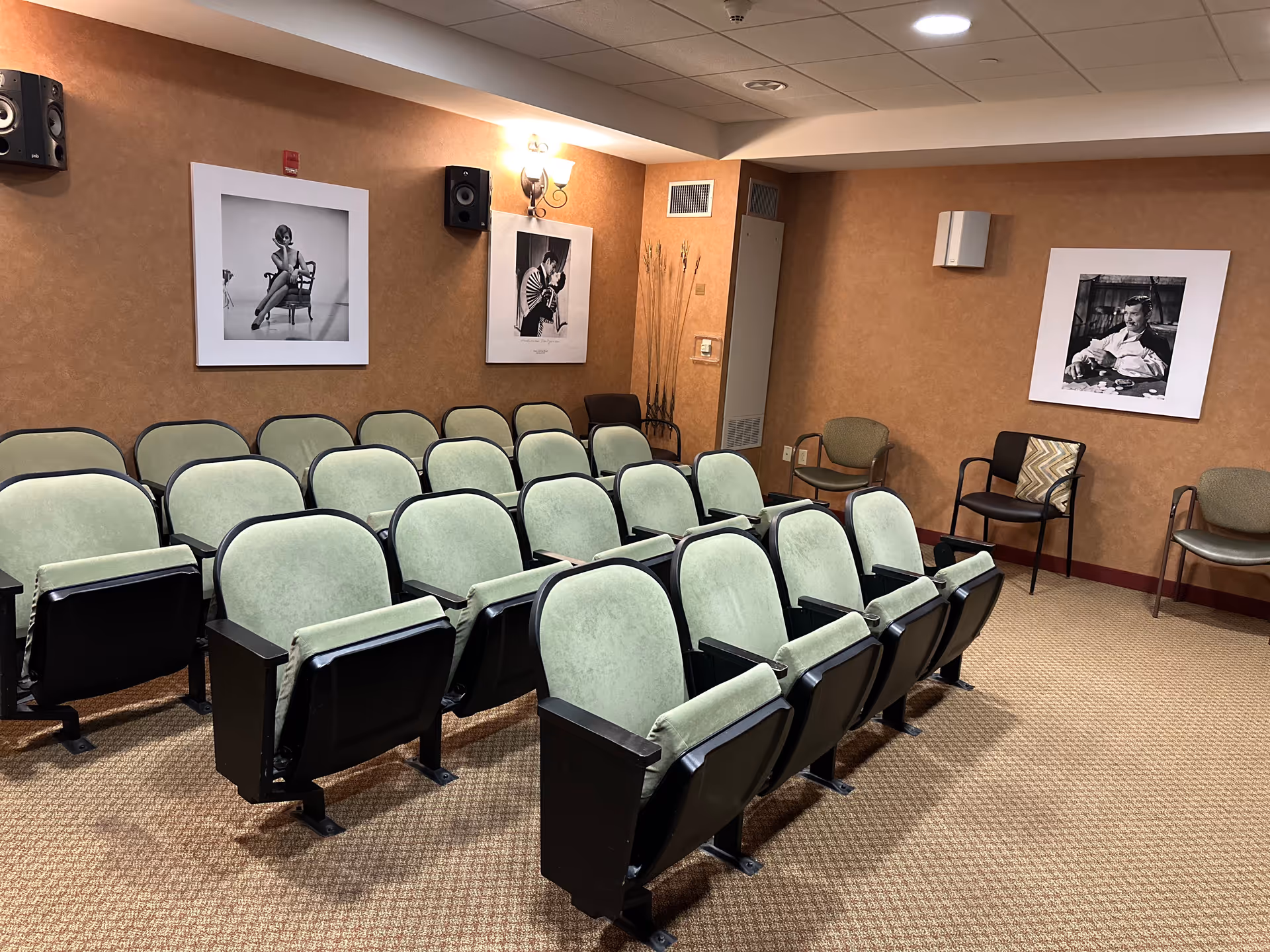 A small theater or presentation room with three rows of green cushioned seats with black armrests. The walls are brown with three black and white framed photographs of people. There are additional chairs along the walls and two wall-mounted speakers. The room has a carpeted floor and ceiling lights.
