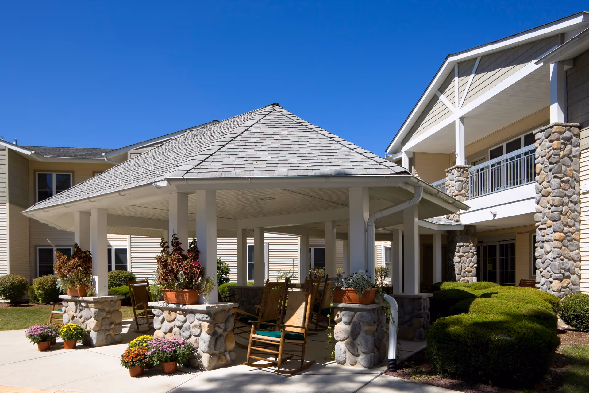 Outdoor covered seating area with a shingled roof supported by white columns on stone bases, surrounded by potted plants and rocking chairs, adjacent to a beige building with stone accents and a balcony under a clear blue sky.