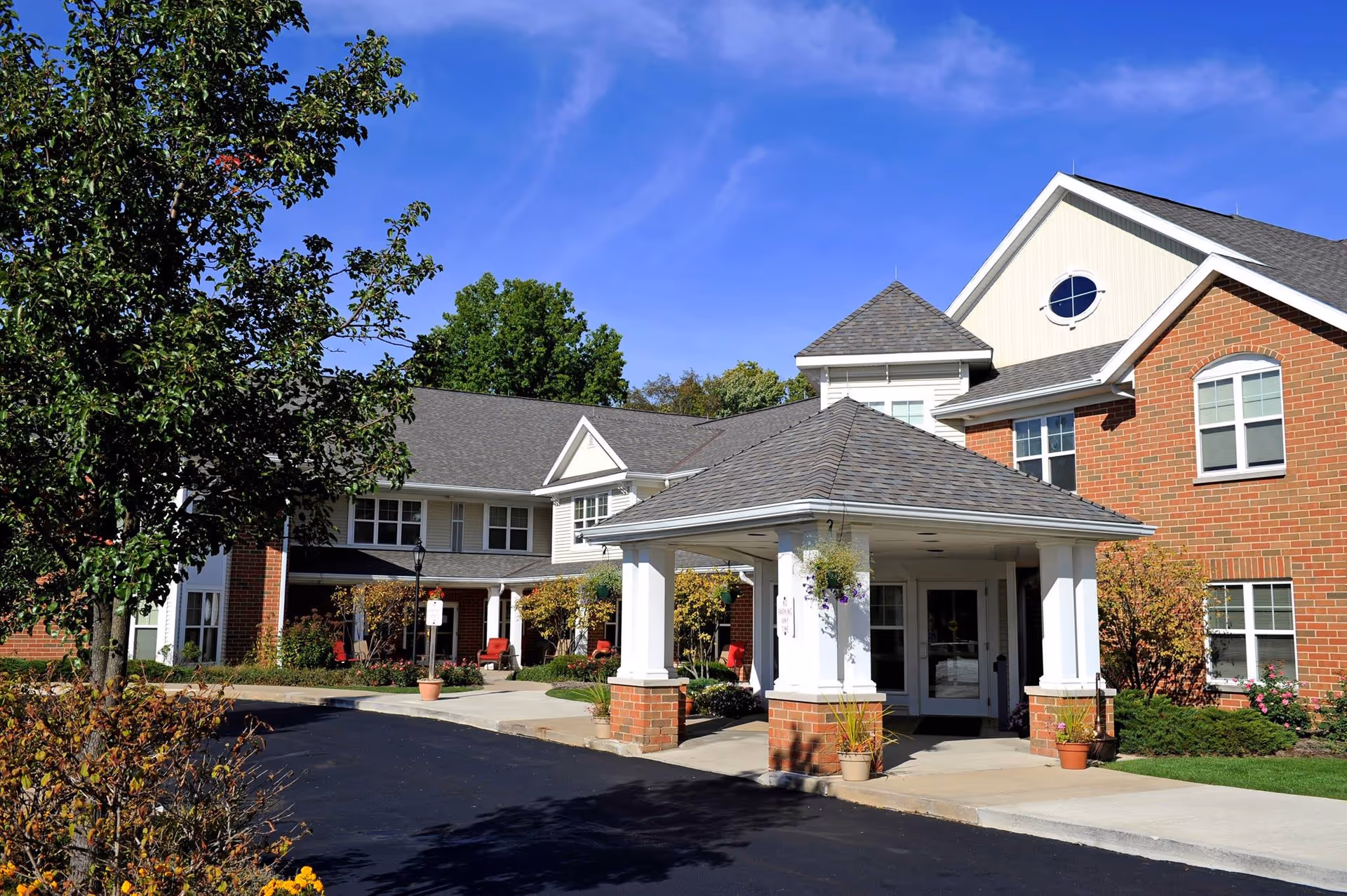 Exterior view of a senior living facility named The Weils, featuring a covered entrance with white columns and brick bases, surrounded by landscaping with trees, shrubs, and potted plants under a clear blue sky.
