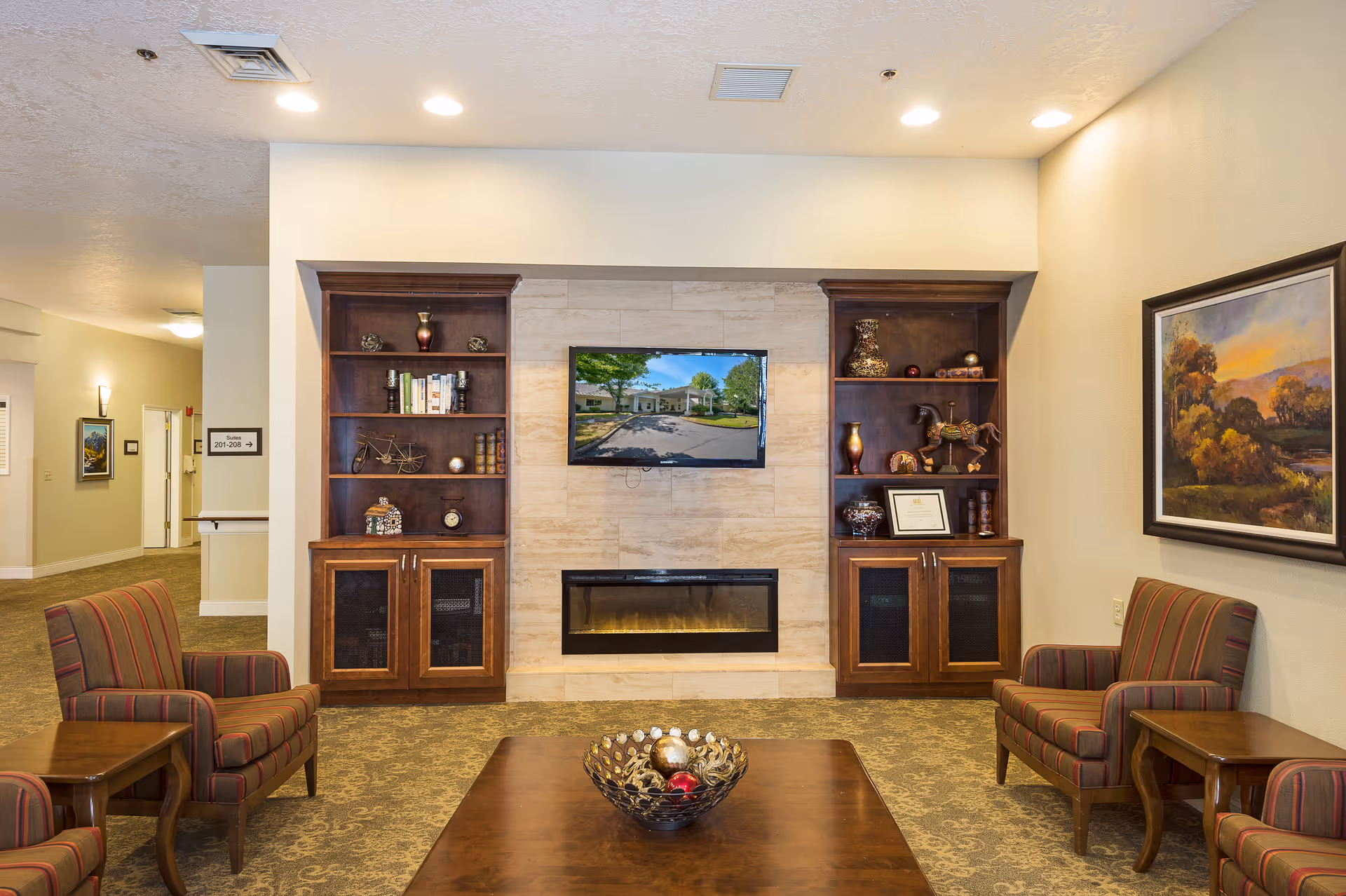 Shared living room with built-in wood shelving and a wall-mounted TV above a fireplace, striped armchairs and a coffee table with a decorative bowl.