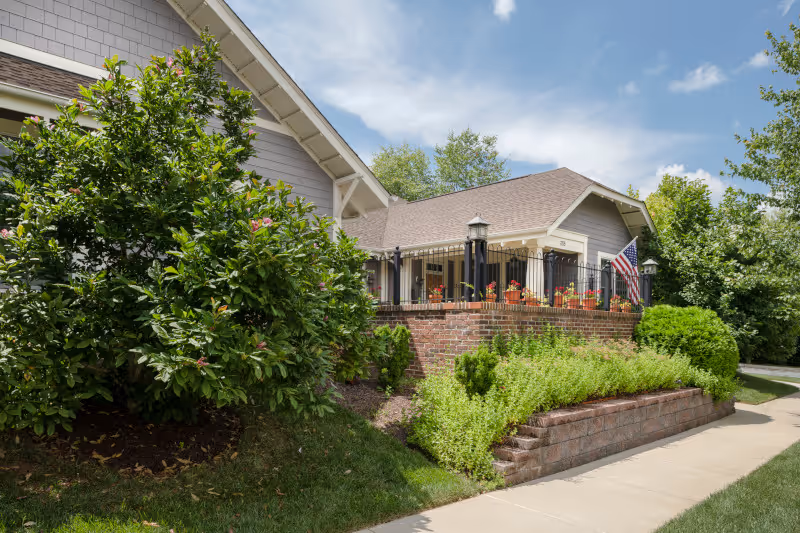 Exterior view of a residential building with a brick raised patio adorned with potted plants and an American flag. The building has light-colored siding and a shingled roof, surrounded by green bushes and trees under a partly cloudy sky.
