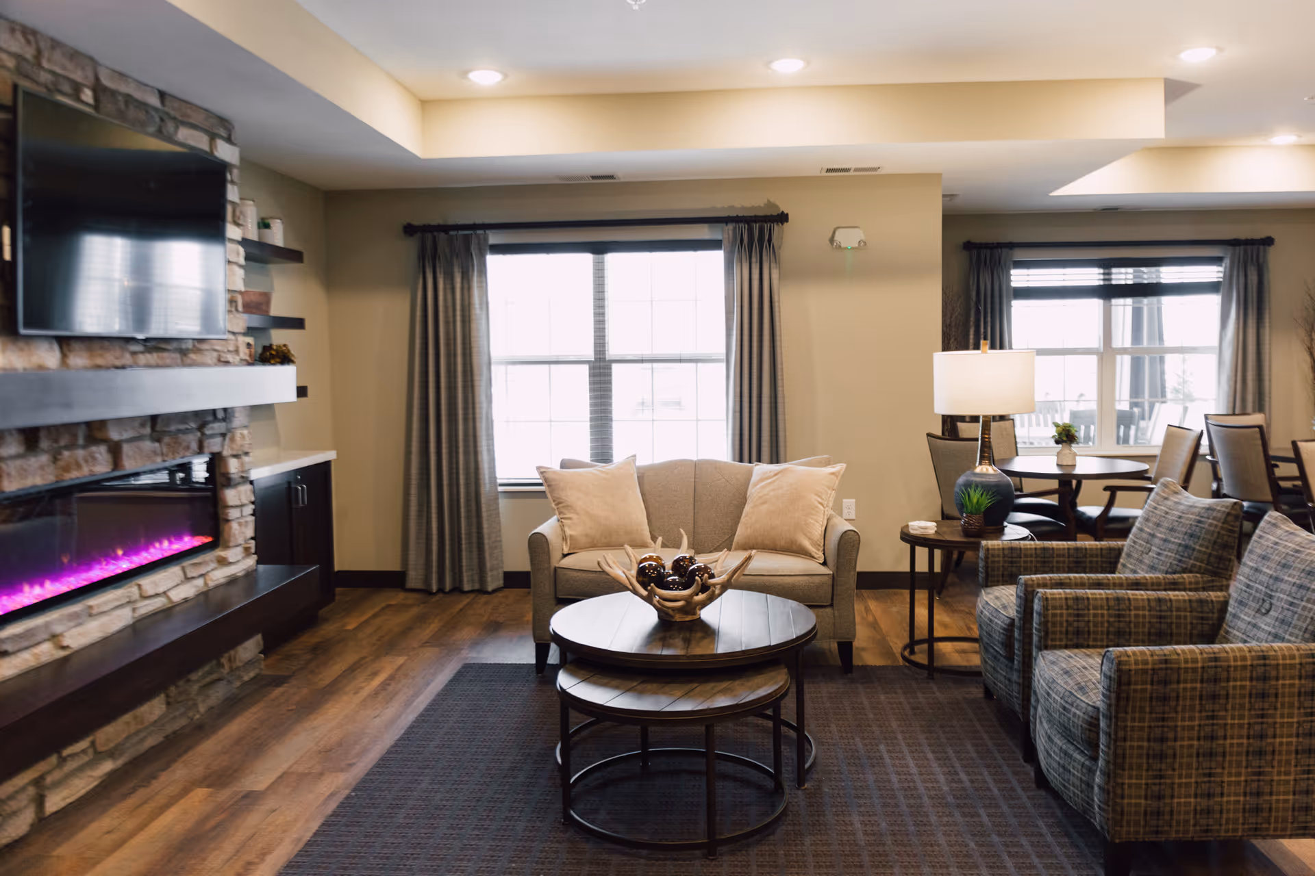 Bright communal living room with sofas, armchairs, round coffee tables, a wall-mounted TV and electric fireplace.