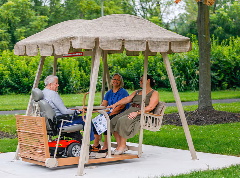 Three people sitting on a covered outdoor swing in a garden area. One person is in a motorized wheelchair, and the other two are seated on the swing bench, smiling and engaging in conversation. The background shows green grass, trees, and shrubs.