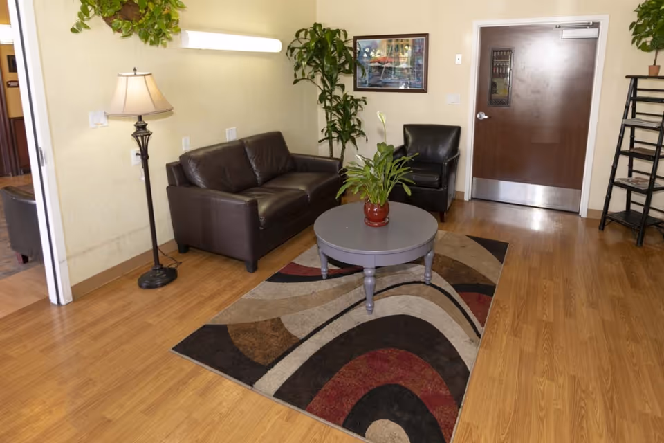 A cozy living room area with a dark brown leather sofa and matching armchair, a round gray coffee table with a potted plant on top, a floor lamp, a colorful patterned rug, a tall green plant in the corner, a framed picture on the wall, and a wooden door with a small window.