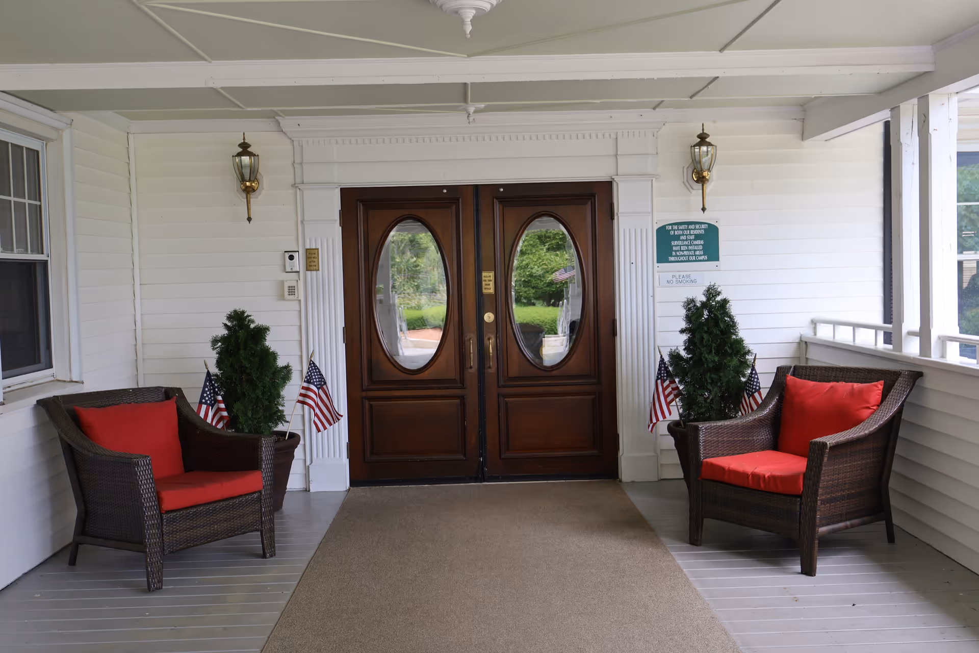 Covered porch area with two dark wicker chairs with red cushions on either side of double wooden doors with oval glass panels. Small potted evergreen plants and American flags are placed next to each chair. The porch has white siding and two wall-mounted lantern-style lights.