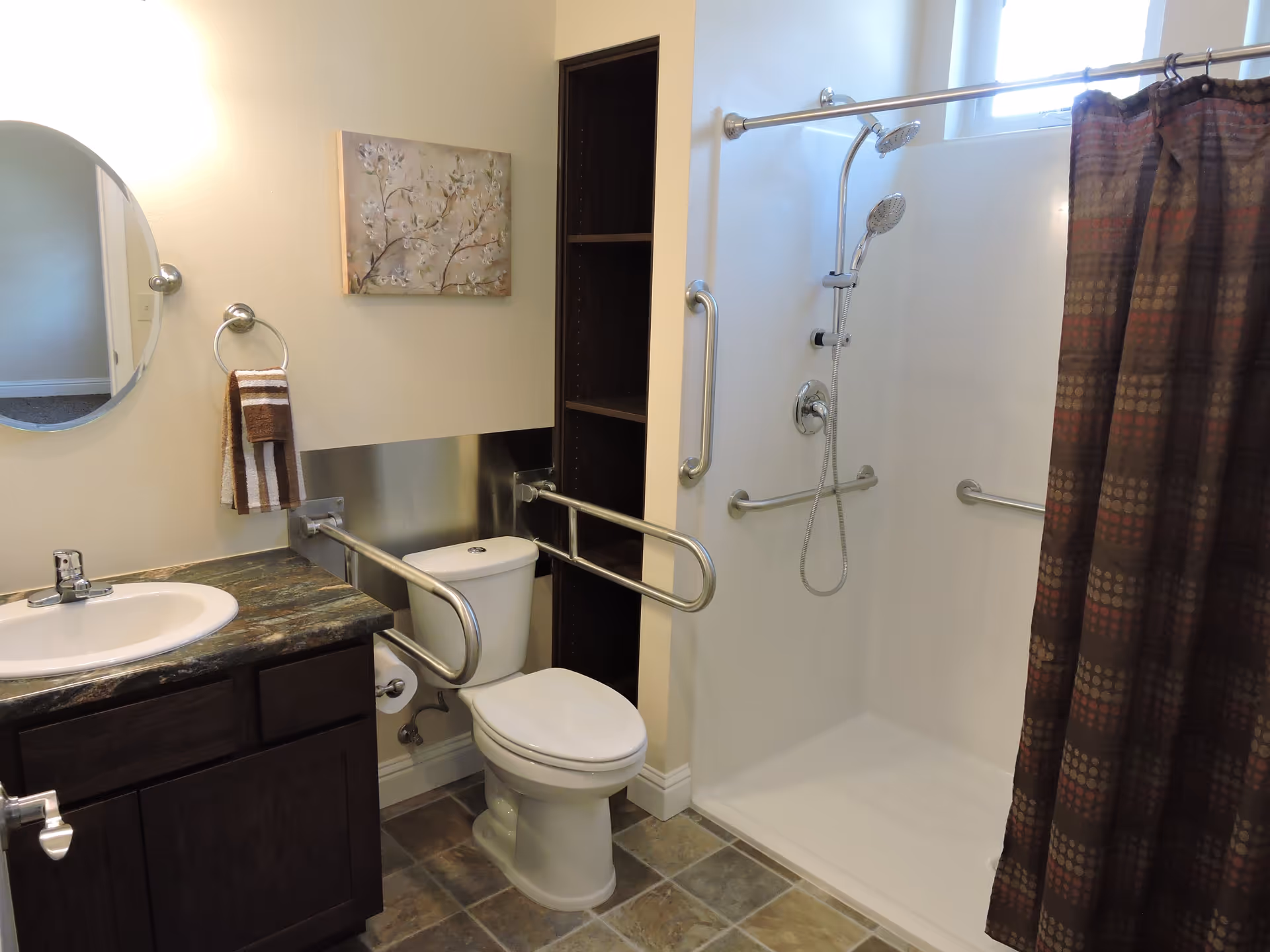 A bathroom with a sink and countertop on the left, a toilet with safety grab bars around it in the center, and a walk-in shower with multiple grab bars and a handheld showerhead on the right. A round mirror and a towel ring with a striped towel are mounted on the wall above the sink. There is a small window above the shower and a decorative painting on the wall above the toilet.