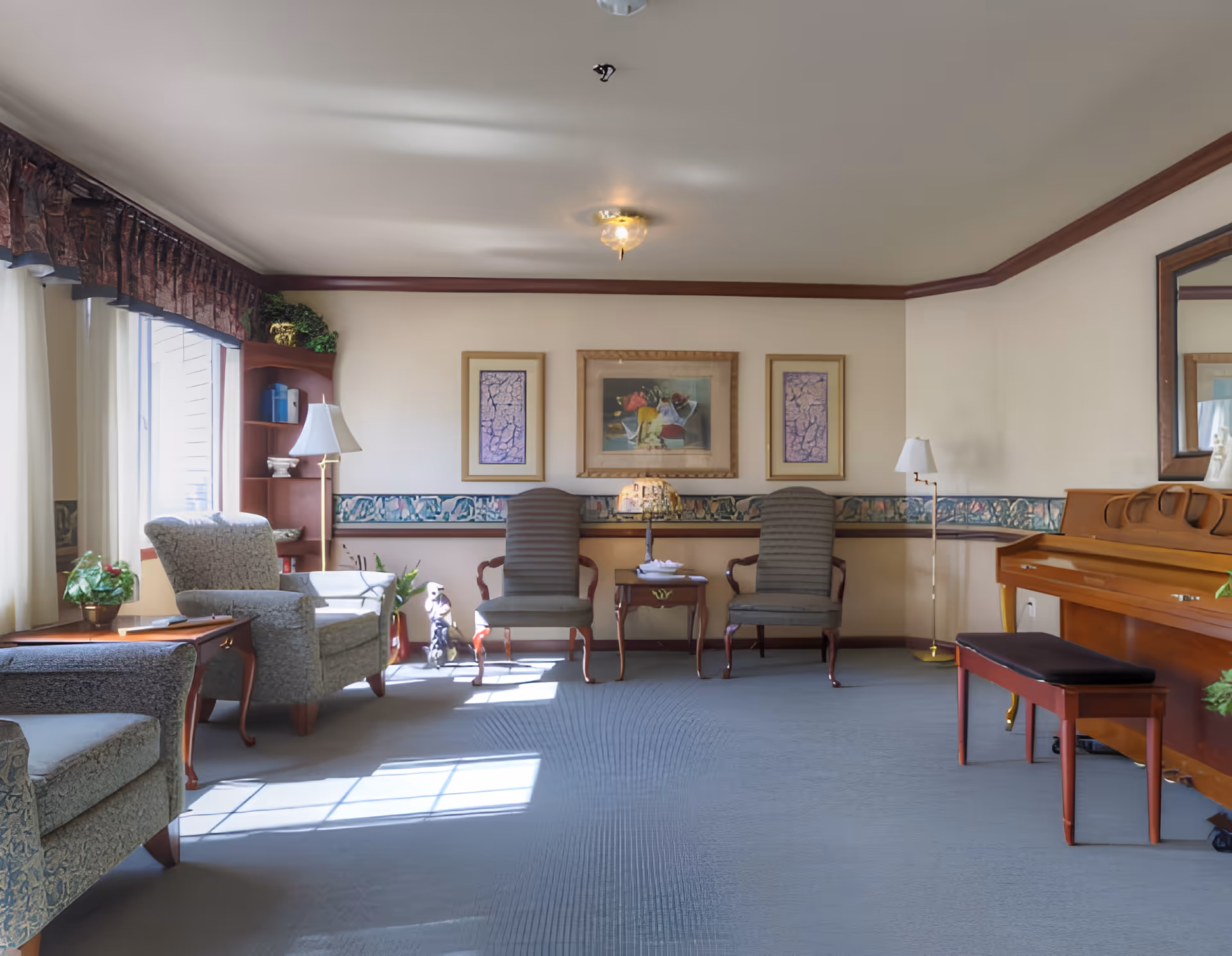 A well-lit living room with large windows on the left letting in natural light. The room features two upholstered armchairs, two wooden chairs with cushions, a small wooden table with a decorative lamp, a piano with a bench on the right, and framed artwork on the wall. There are plants and a bookshelf in the corner, and the room has a calm, inviting atmosphere.