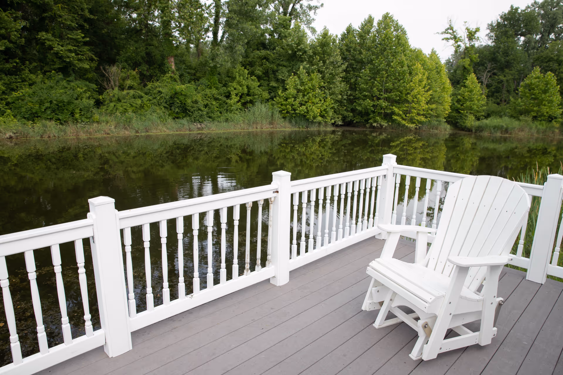 A white wooden Adirondack chair on a gray deck with white railing overlooking a calm pond surrounded by green trees and foliage.