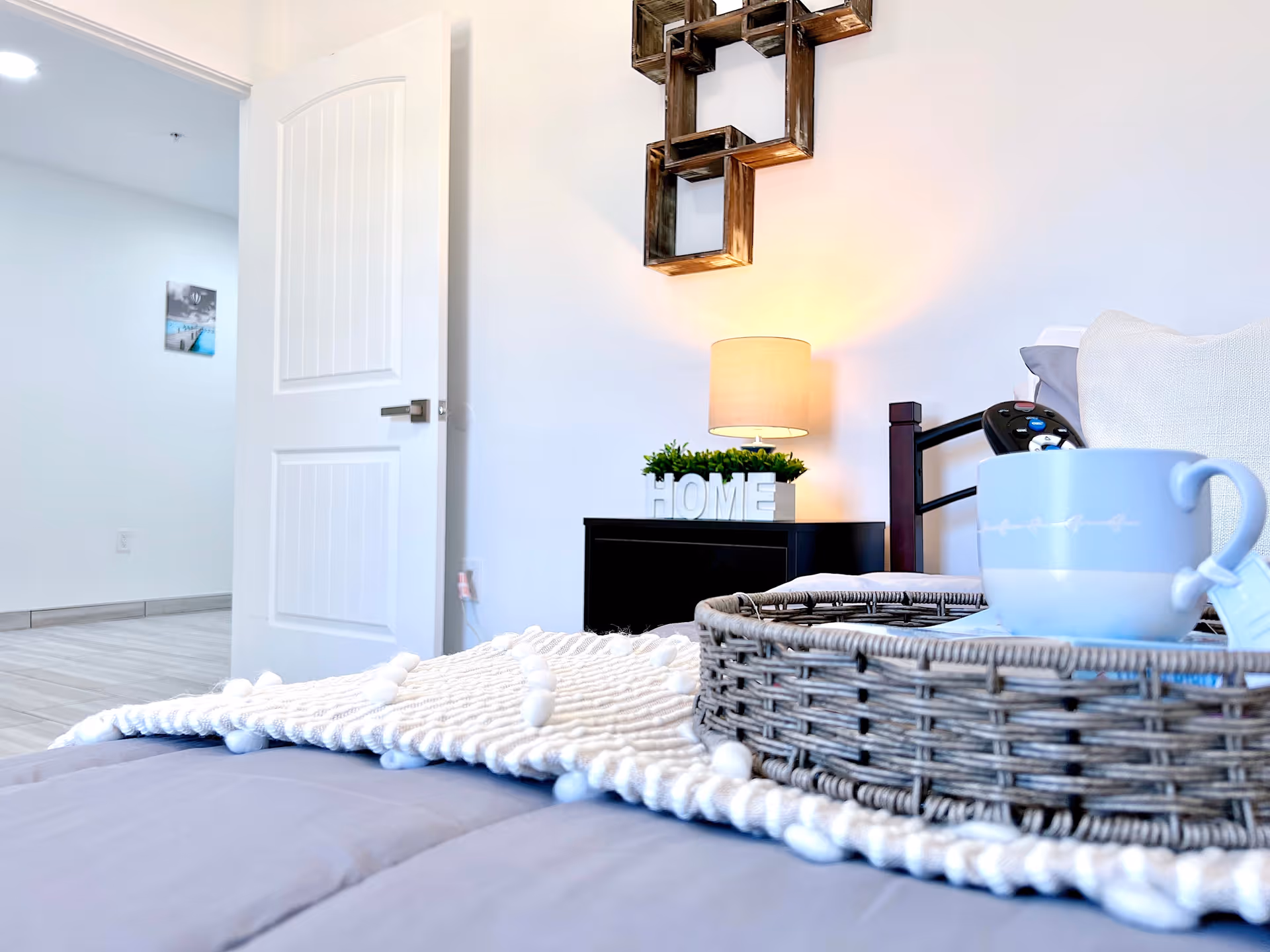 A cozy bedroom scene with a bed covered in a gray comforter and a white textured throw blanket. On the bed is a wicker tray holding a light blue cup. In the background, there is a black nightstand with a small lamp, a decorative plant, and white letters spelling 'HOME'. A modern wooden wall decoration hangs above the nightstand. A white door is open, leading to another room with light-colored flooring and a wall picture.