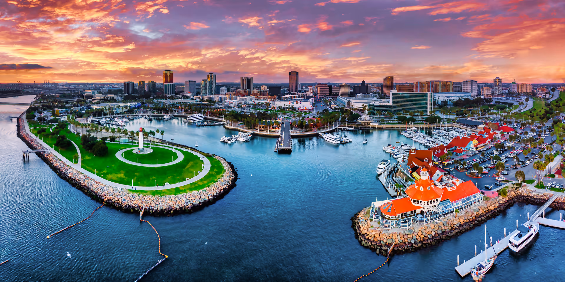 Aerial view of a coastal city harbor at sunset with a vibrant sky. The image shows a curved green park with a lighthouse on the left, a marina with boats docked, and buildings with red roofs on the right. The city skyline is visible in the background with various high-rise buildings.