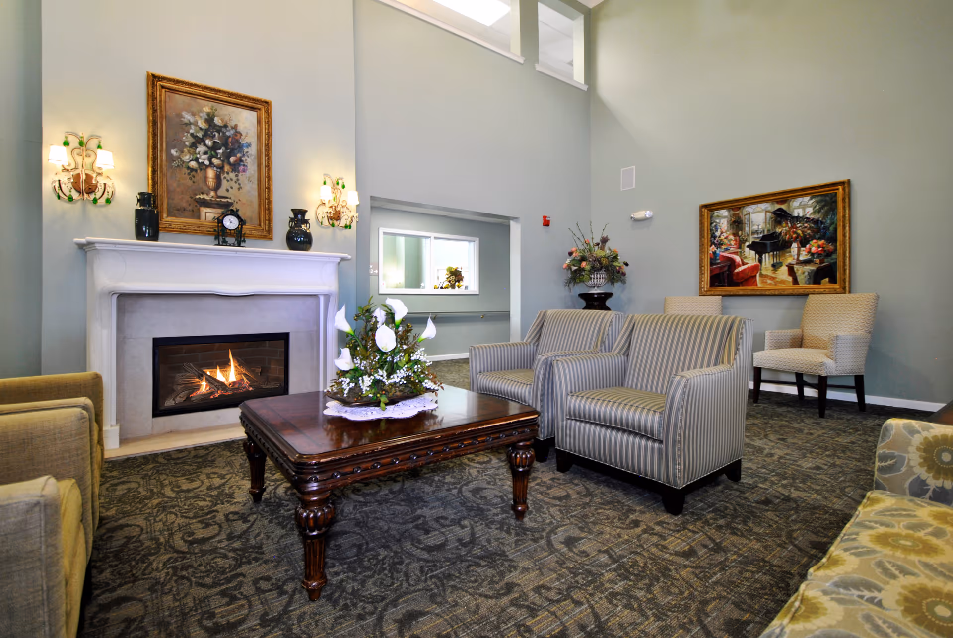 A cozy living room area with a lit fireplace, a wooden coffee table adorned with a floral arrangement, two striped armchairs, a beige armchair, and a floral-patterned sofa. The walls are painted light green and decorated with two framed paintings and wall sconces with lights.