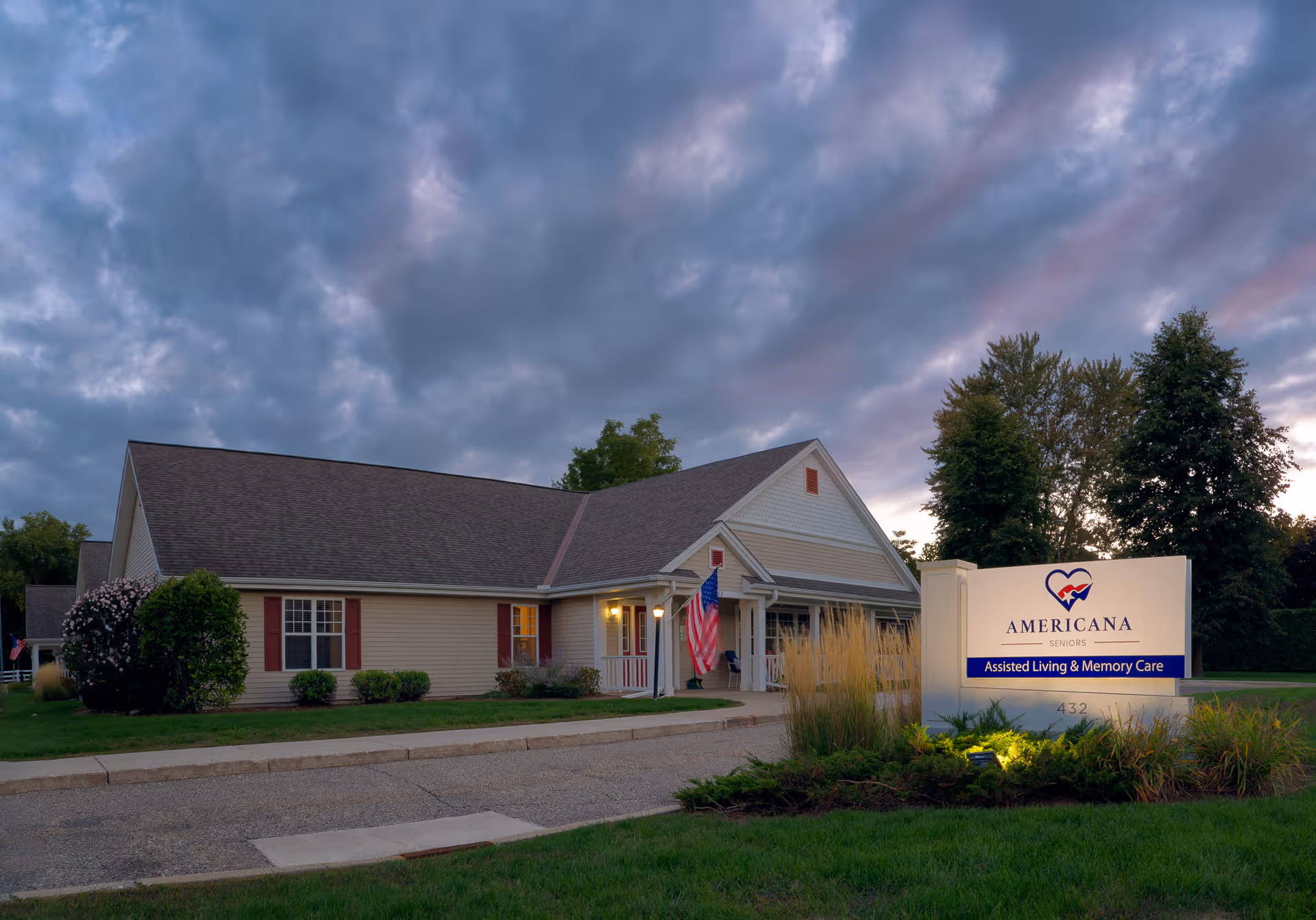Single-story senior living facility with a front entrance, lawn and illuminated sign for Americana Seniors under a dramatic cloudy sky.