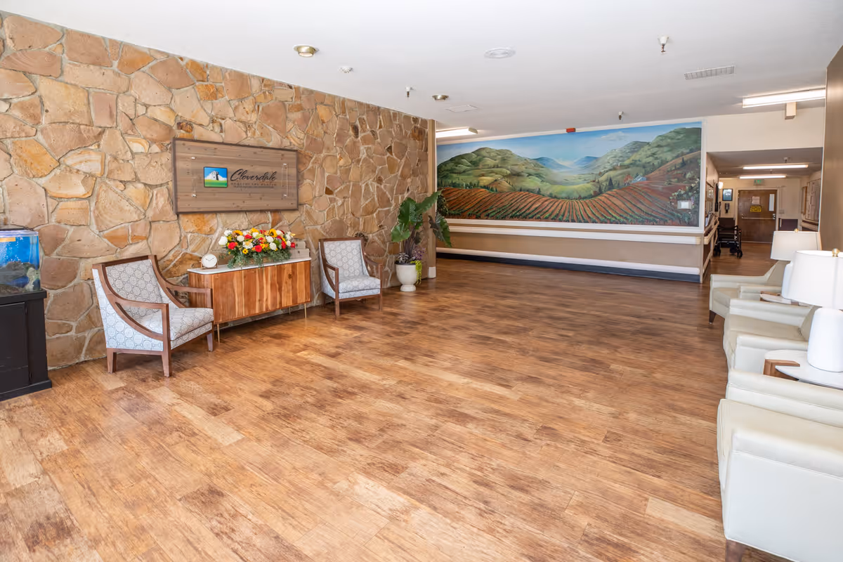 A spacious interior common area with wooden flooring and a stone accent wall featuring a Cloverdale Healthcare Center sign. The room has several chairs arranged along the walls, a small cabinet with a flower arrangement, a fish tank, and a large mural depicting a scenic landscape of hills and fields.