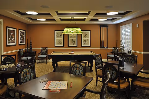 A well-lit game room with a pool table in the center, surrounded by several tables and chairs. One table has a Scrabble board game set up. The walls are decorated with framed artwork, and there is a window with white shutters on the right side. The ceiling features recessed lighting and a decorative light fixture above the pool table.