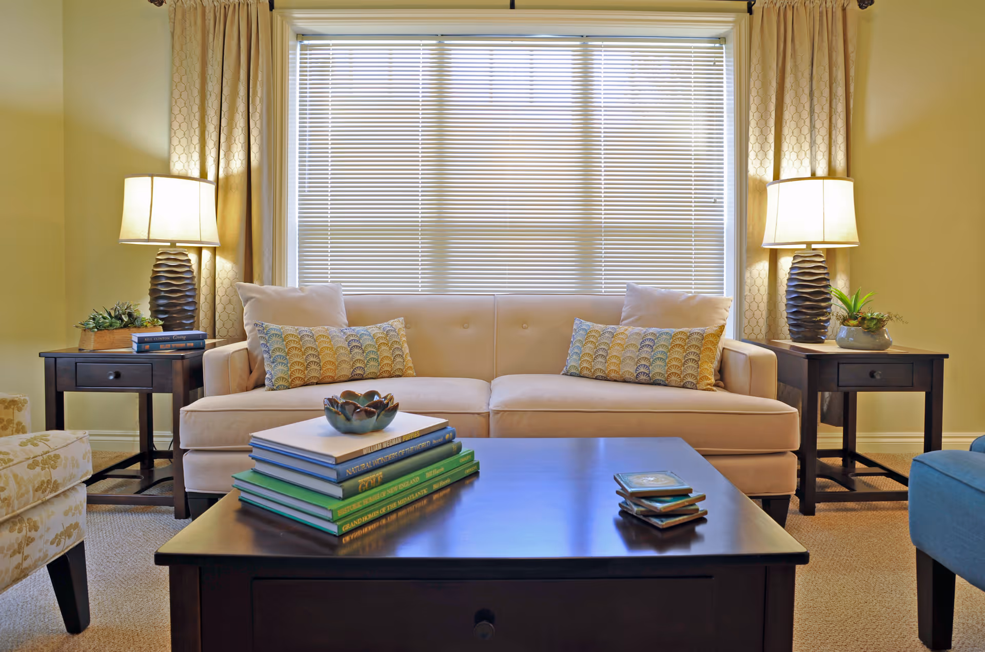 Sunlit living room with a beige sofa, patterned pillows, two side tables with lamps, and a coffee table topped with stacked books.