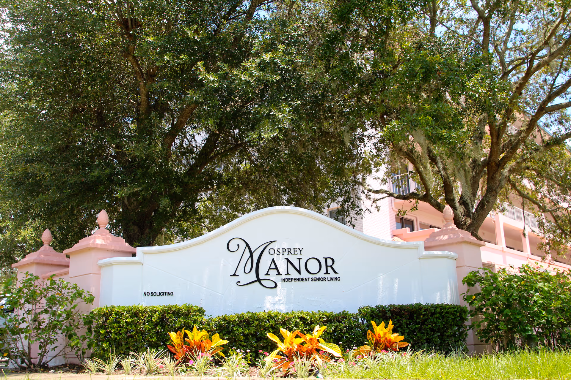Entrance sign for Osprey Manor Independent Senior Living surrounded by landscaping and large oak trees with the building visible behind.