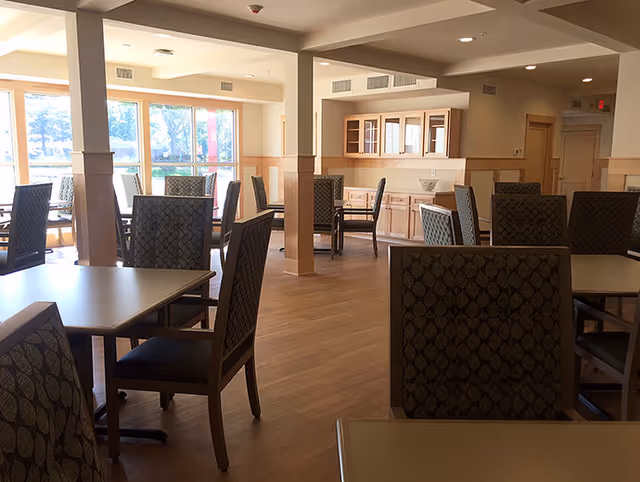Bright communal dining room with multiple tables and upholstered chairs, wooden floors, built-in cabinets, and large windows letting in daylight.