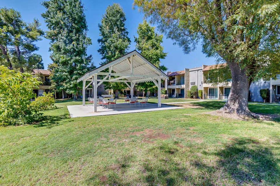 Outdoor view of a senior living facility courtyard with a white covered pavilion featuring tables and chairs. Surrounding the pavilion are green lawns, large trees, and two-story residential buildings with balconies and patios under a clear blue sky.