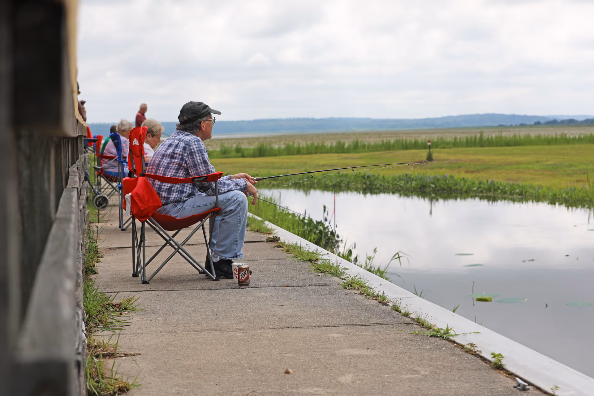 Several elderly people sitting on folding chairs along a concrete path beside a calm body of water, fishing. The scene is outdoors with grassy fields and a cloudy sky in the background.