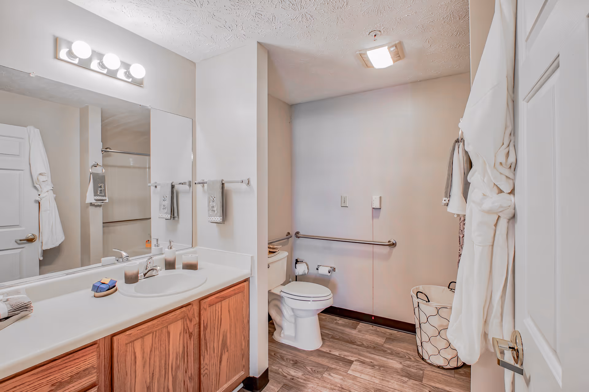 A clean and well-lit bathroom with a large mirror above a sink with wooden cabinetry. There are three lit bulbs above the mirror, a white toilet with grab bars on the wall, a laundry basket, and white robes hanging on the door and wall. The floor has wood-like tiles.