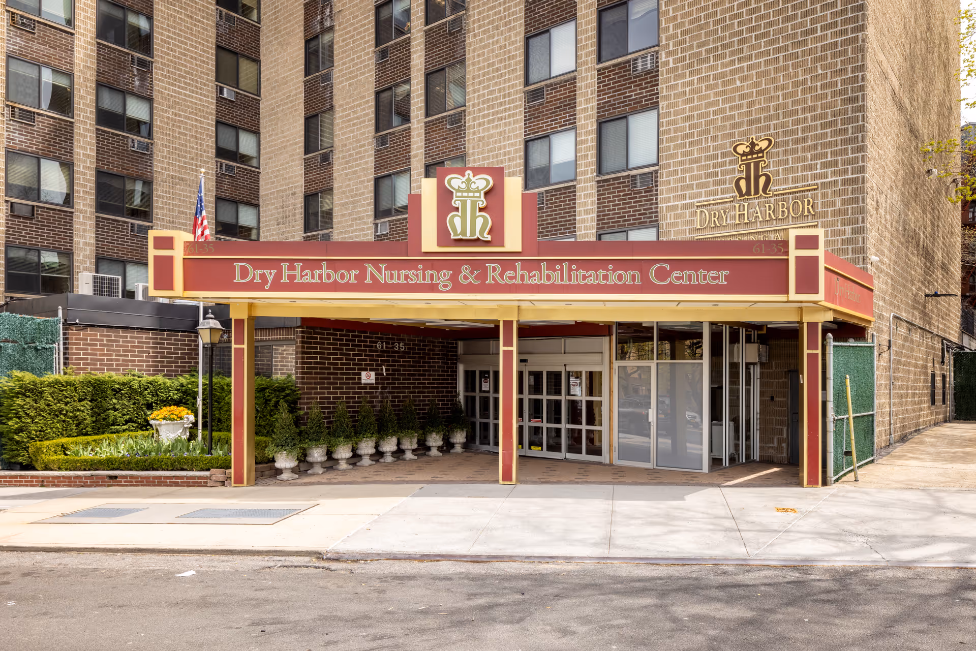 Entrance of Dry Harbor Nursing & Rehabilitation Center with a red and gold canopy displaying the facility name. The building is a multi-story brick structure with several windows. There are potted plants and greenery near the entrance, and an American flag is visible on the left side.