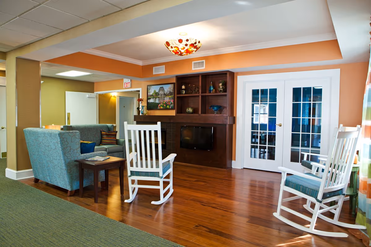 A cozy living room area with two white rocking chairs with blue cushions, a blue patterned armchair, and a gray sofa with colorful pillows. The room has wooden flooring, an orange accent wall with a built-in dark wood shelving unit and a small TV. There is a framed picture above the shelving unit and double glass doors on the right side. The ceiling has a decorative light fixture and there is a green carpeted area on the left side.