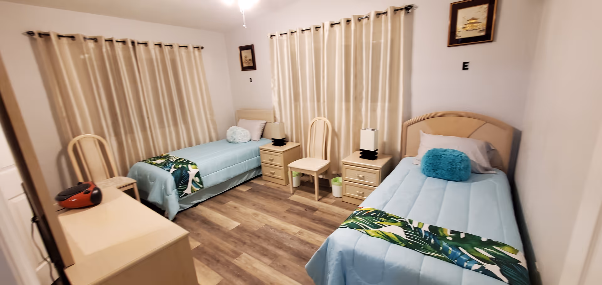 A bedroom with two single beds, each with light blue bedding and decorative pillows. Between the beds are two nightstands with lamps and a chair. The room has wood flooring, beige curtains covering the windows, and light-colored walls with framed pictures.