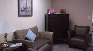 A cozy living room area with a brown sofa adorned with a blue pillow, a brown armchair, a wooden cabinet with books and decorative items on top, a table lamp with a white shade, and a framed picture on the wall.