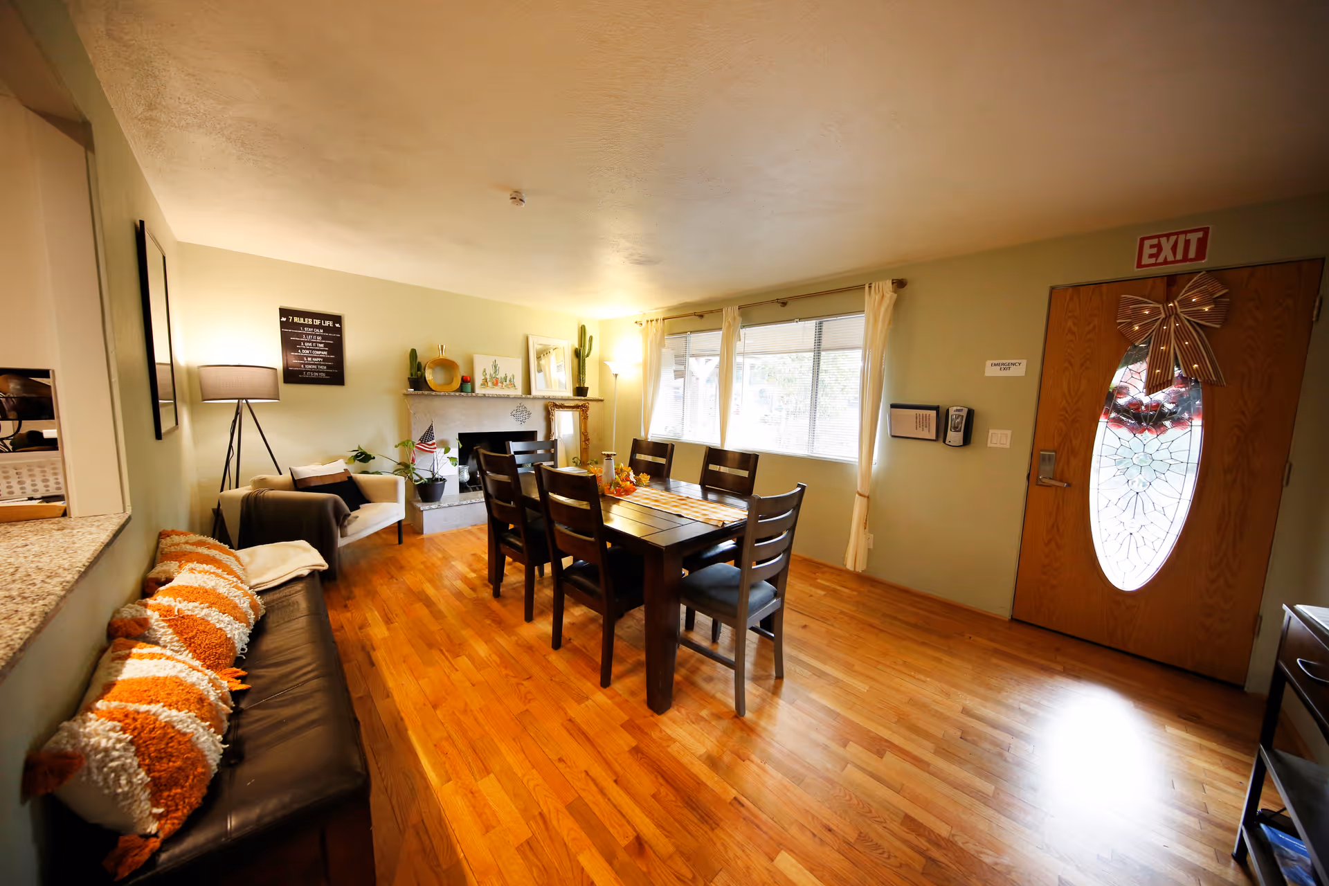 A warm and inviting dining area with a wooden dining table and six chairs in the center. To the left, there is a black leather bench with orange and white striped pillows, and a cozy armchair next to a floor lamp. The room features hardwood floors, a fireplace decorated with plants and framed pictures, and a large window with white curtains letting in natural light. A wooden front door with an oval glass panel and a decorative bow is visible on the right side, along with an exit sign above it.